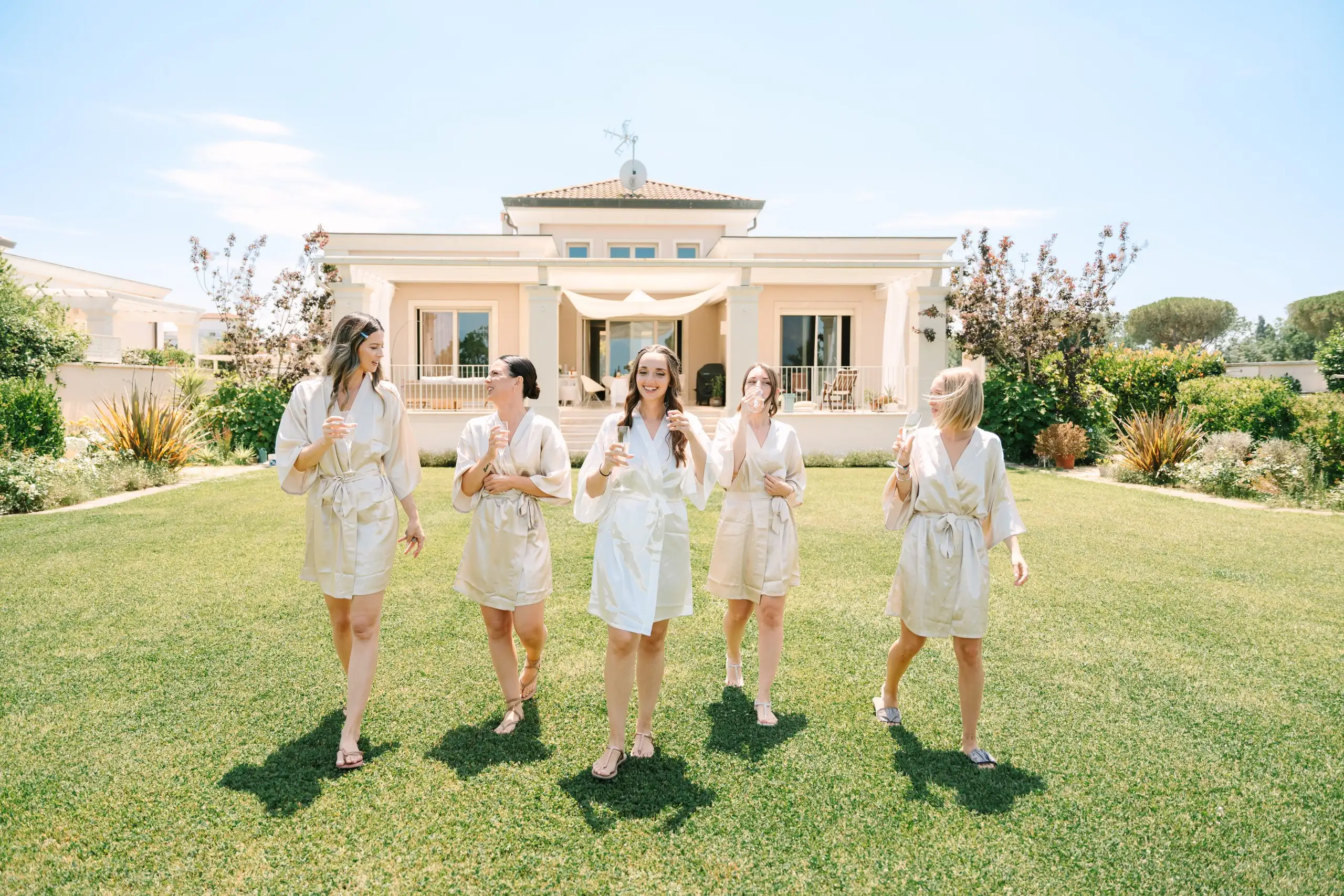 Bride and bridesmaids in matching satin robes on the lawn before a villa