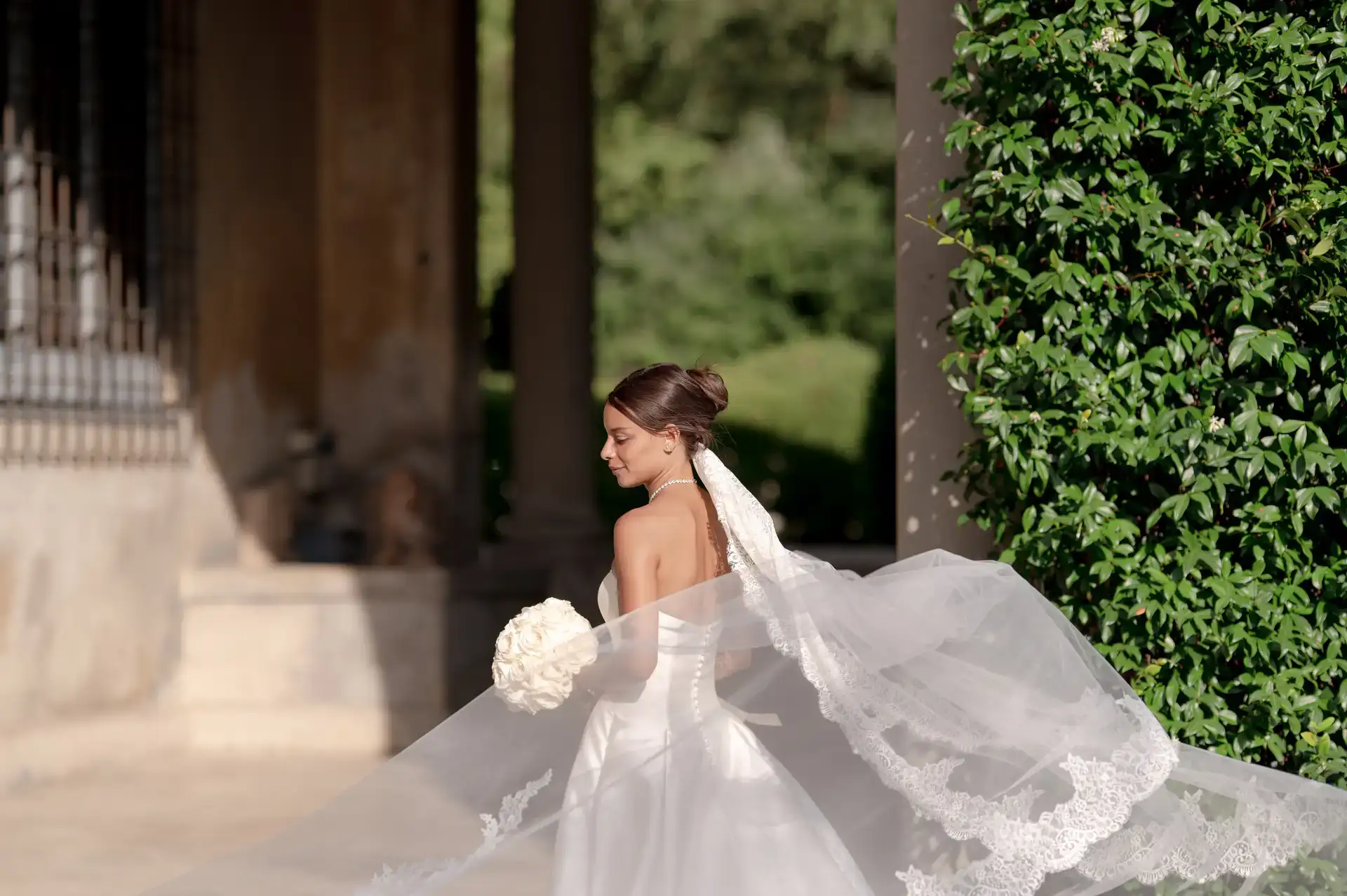 Bride with a flowing lace veil and white bouquet in a villa garden