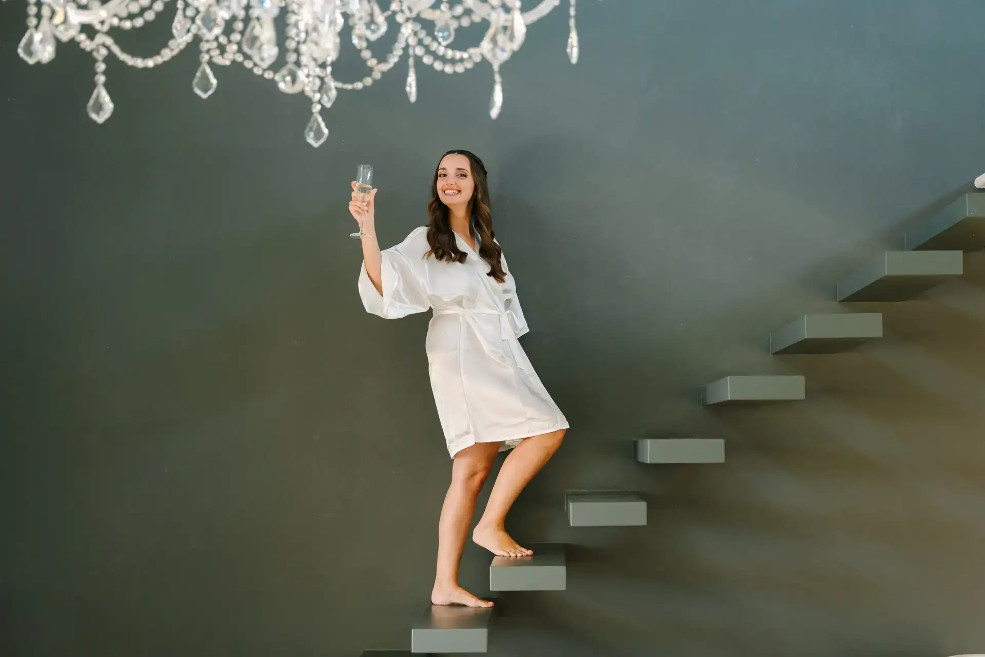 Bride in a white silk robe with a glass of champagne under a crystal chandelier